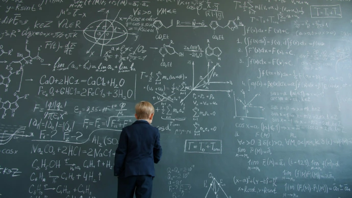 Young boy in suit studies complex math formulas on blackboard.